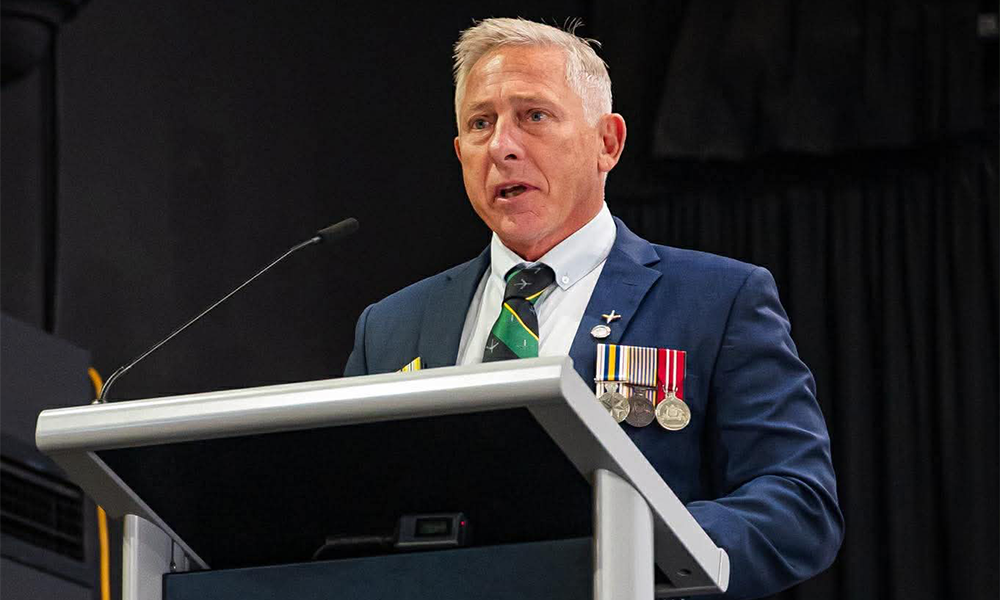 Will Collett speaking at a lectern in a suit with service medals
