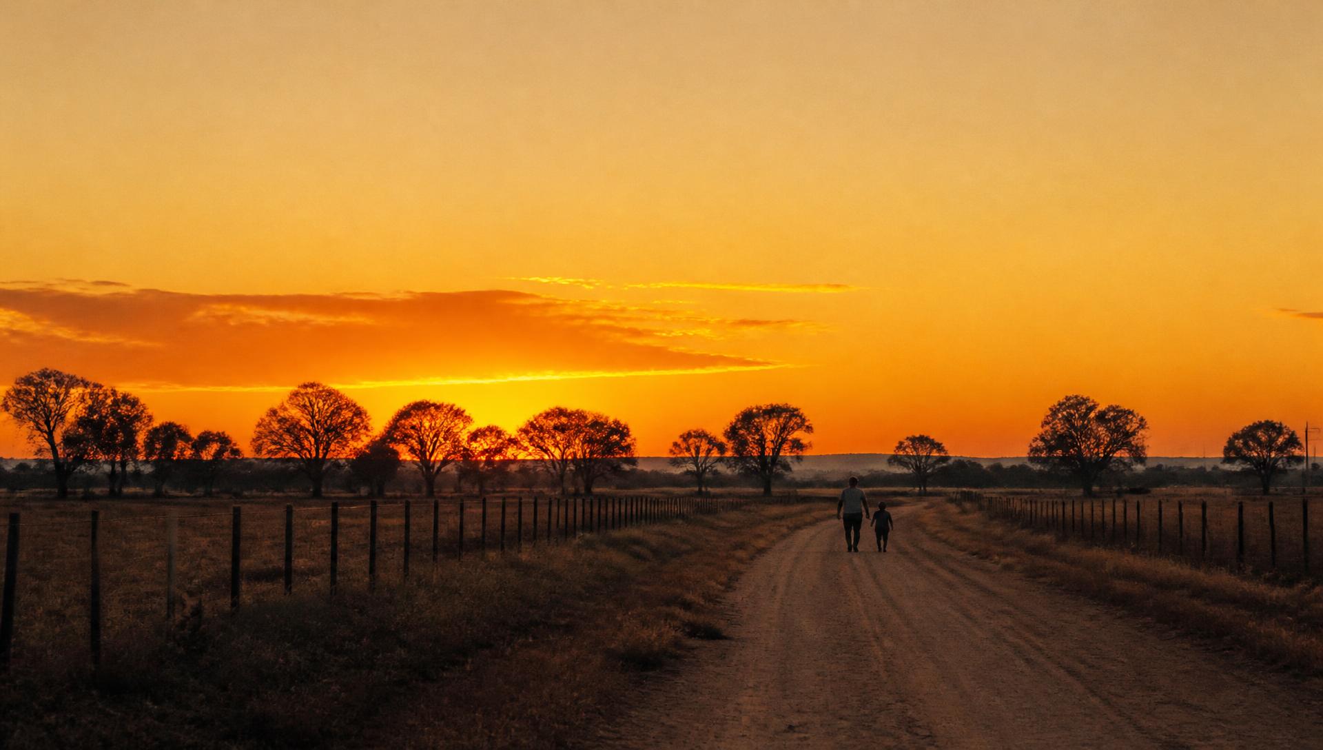 A man and a young boy walking a dirt farm road at sunset in rural Australia