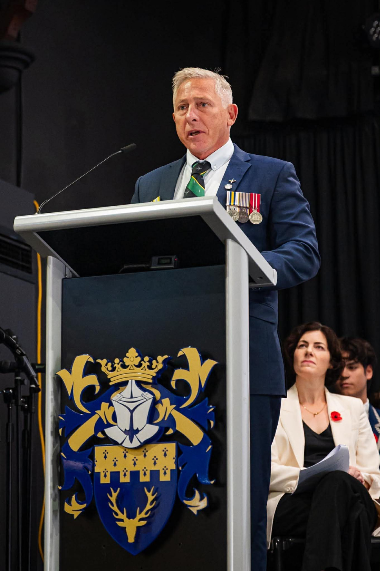 Will Collett speaking at a lectern in a suit with service medals