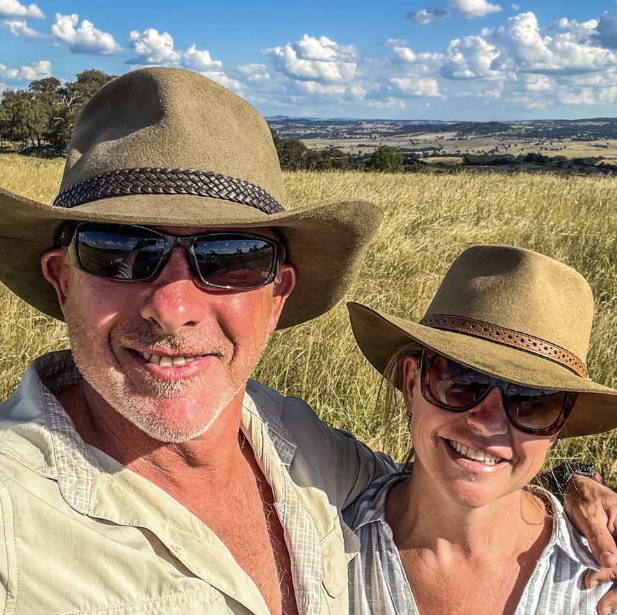 Will Collett and a woman wearing broad-brimmed hats in long grass, with rolling farmland behind them